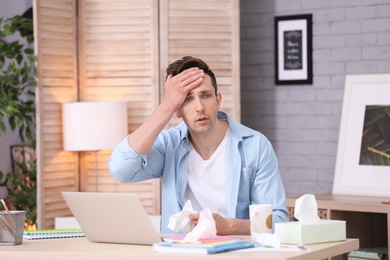 Photo of Sad exhausted man with tissue suffering from cold while working with laptop at table