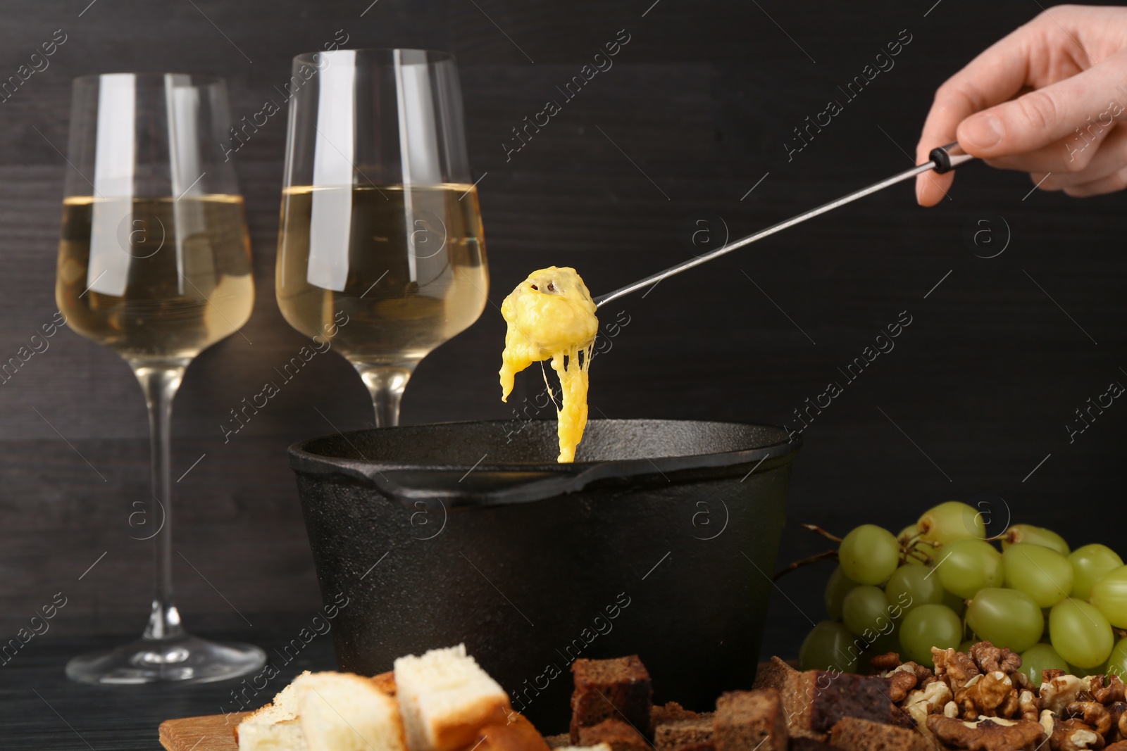 Photo of Woman dipping piece of bread into fondue pot with melted cheese at black wooden table, closeup