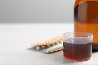 Photo of Measuring cup with syrup, pills on white table, closeup and space for text. Cold medicine