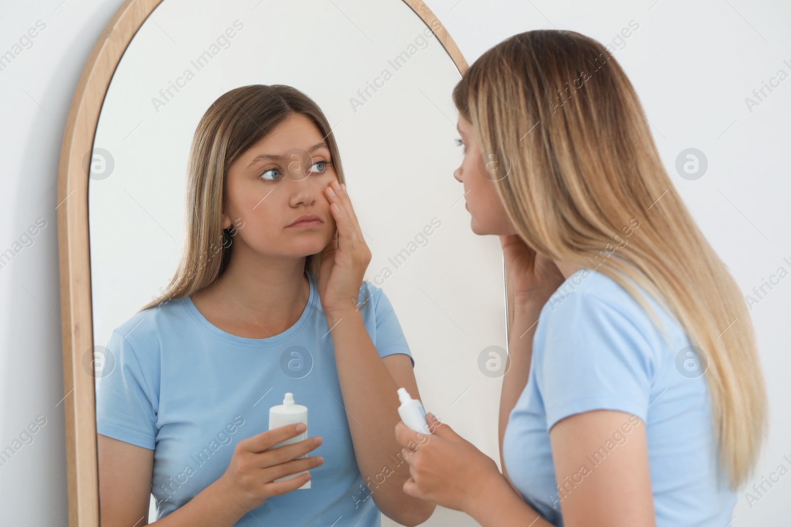 Photo of Sleep deprived young woman covering up dark circles with concealer near mirror at home