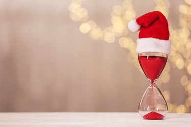 Photo of Hourglass with Santa hat on white table against blurred lights, space for text. Christmas countdown