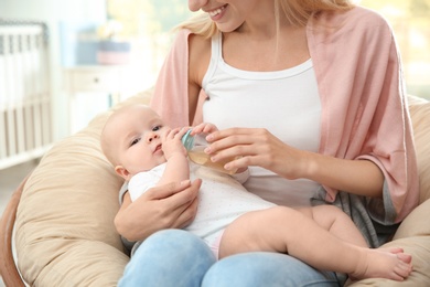 Lovely mother giving her baby drink from bottle in room, closeup