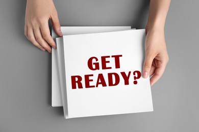 Image of Woman holding paper sheet with text Get Ready? at grey table, top view