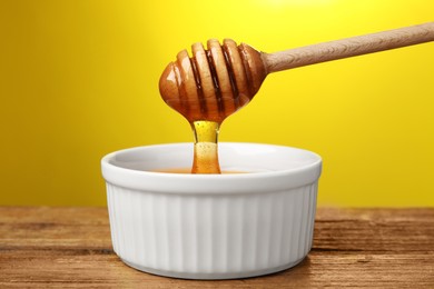 Pouring honey from dipper into bowl at wooden table against yellow background, closeup