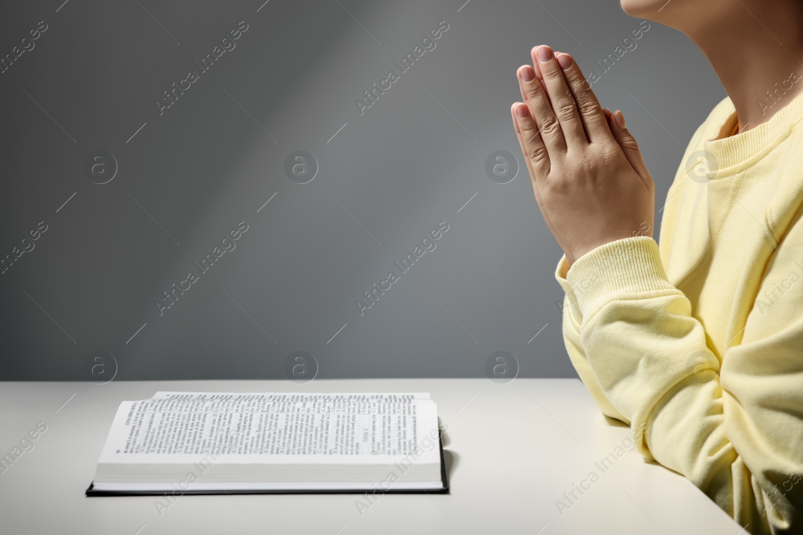 Photo of Woman praying over Bible at white table against grey background, closeup. Space for text