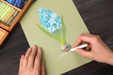 Photo of Woman drawing blue blooming hyacinth on paper with soft pastels at wooden table, closeup
