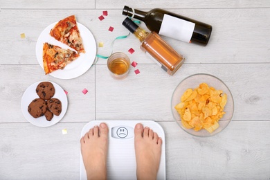 Photo of Woman weighing on scales surrounded by junk food and alcohol after party indoors, top view