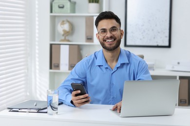 Photo of Happy young man using smartphone while working with laptop at white table in office