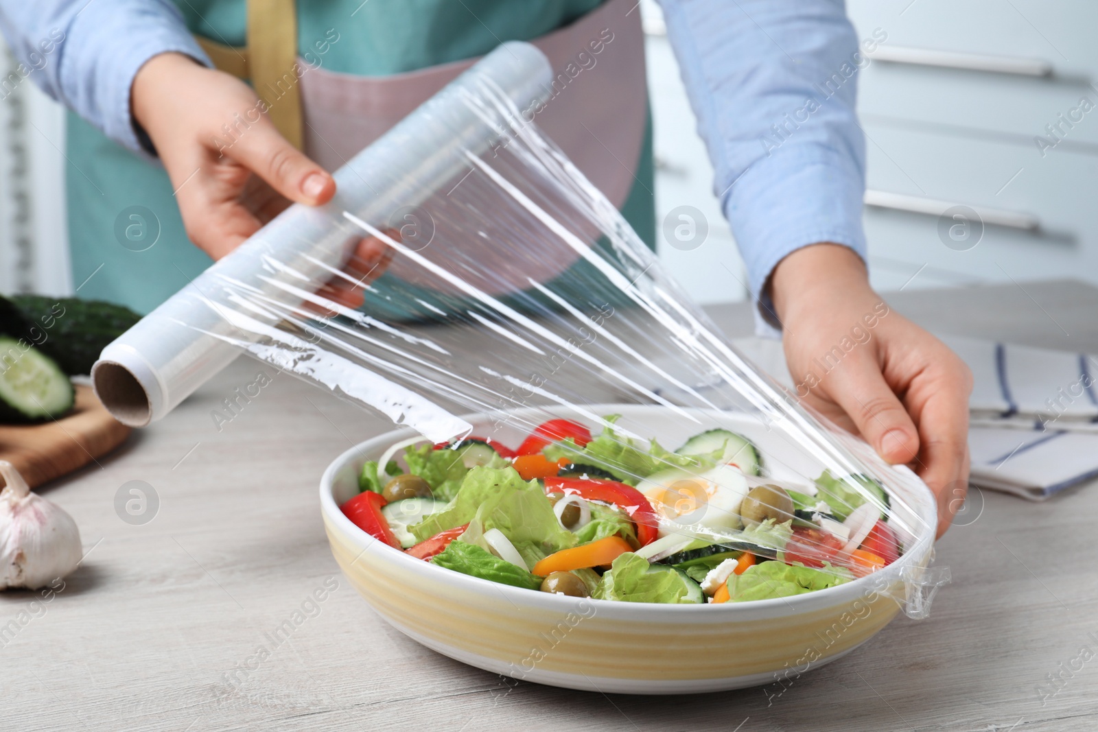 Photo of Woman putting plastic food wrap over bowl of fresh salad at wooden table indoors, closeup