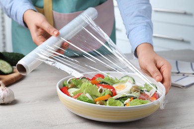 Photo of Woman putting plastic food wrap over bowl of fresh salad at wooden table indoors, closeup