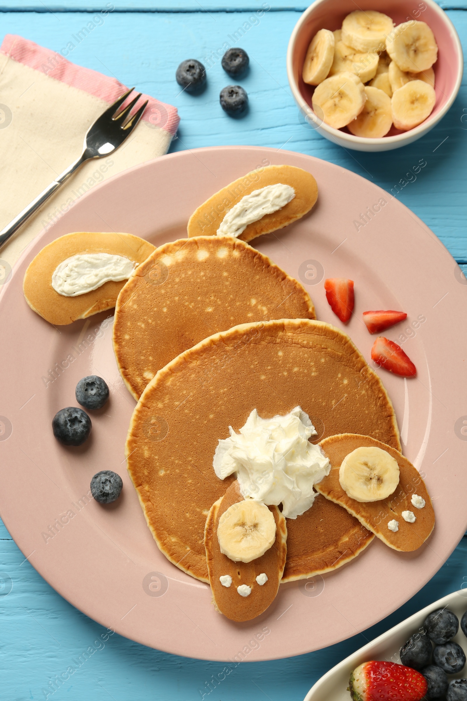 Photo of Creative serving for kids. Plate with cute bunny made of pancakes, berries, cream and banana on light blue wooden table, flat lay