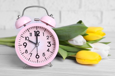 Photo of Pink alarm clock and beautiful tulips on white wooden table against brick wall, closeup. Spring time