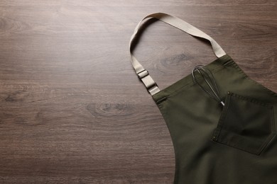 Stylish dark olive apron and whisk on wooden table, top view. Space for text