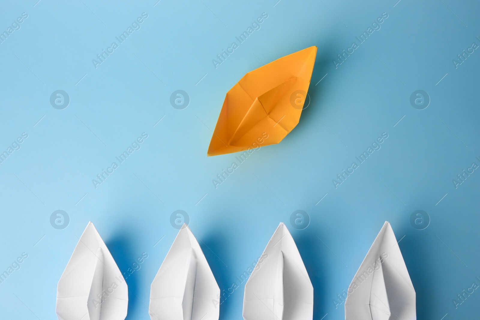 Photo of Yellow paper boat floating away from others on light blue background, flat lay. Uniqueness concept