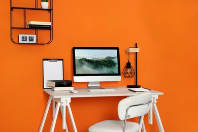 Photo of Workplace with modern computer on wooden desk and comfortable chair near orange wall. Home office