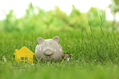 Photo of Piggy bank, house figure and jar with coins on green grass in park. Space for text