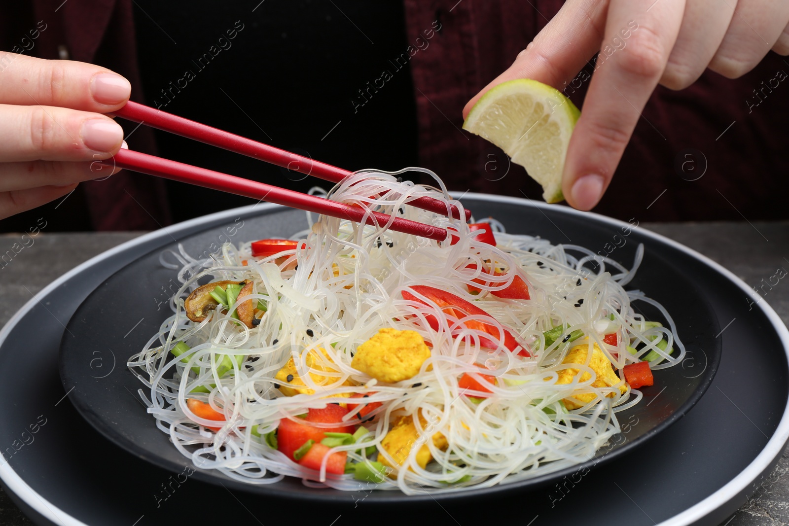 Photo of Stir-fry. Woman squeezing lime into tasty rice noodles with meat and vegetables at table, closeup