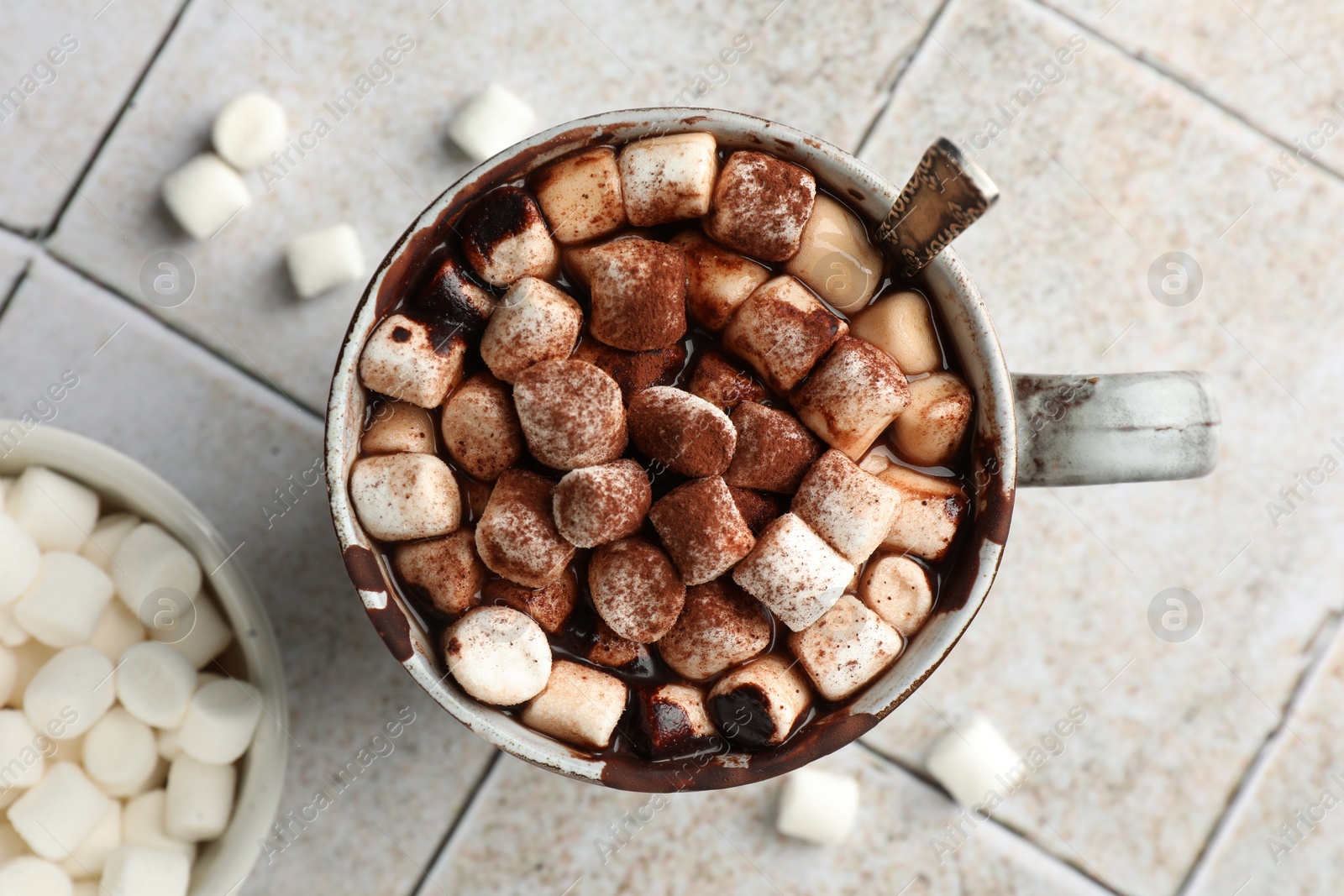 Photo of Delicious hot chocolate with marshmallows, cocoa powder and spoon in cup on tiled table, top view