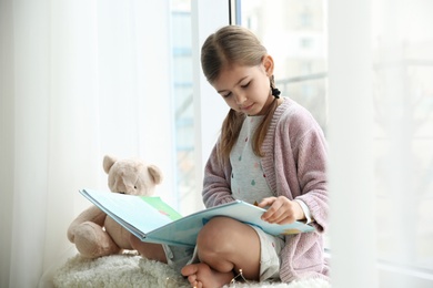 Photo of Cute little girl reading book near window at home