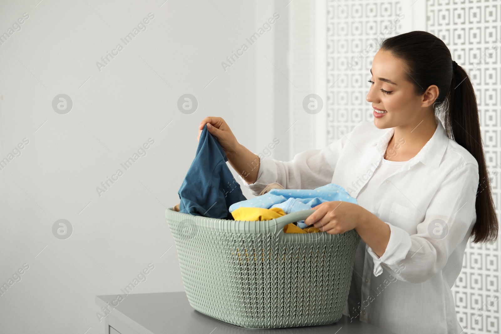 Photo of Young woman with basket full of clean laundry at table indoors