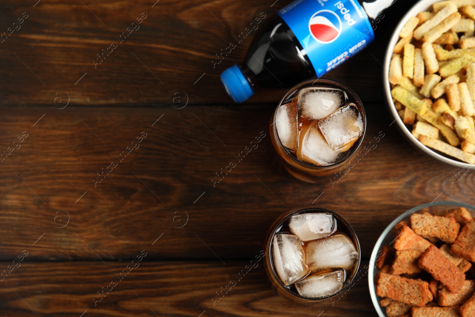 Photo of MYKOLAIV, UKRAINE - FEBRUARY 15, 2021: Glasses and bottle of Pepsi with snacks on wooden table, flat lay. Space for text