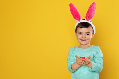 Little boy in bunny ears headband holding Easter egg on color background, space for text