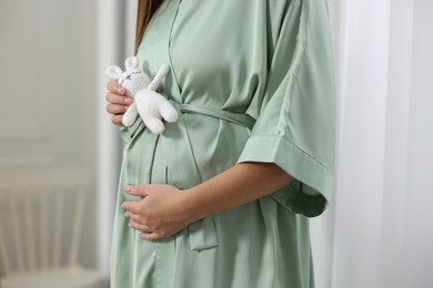 Photo of Pregnant woman in green dressing gown with bunny toy near window indoors, closeup