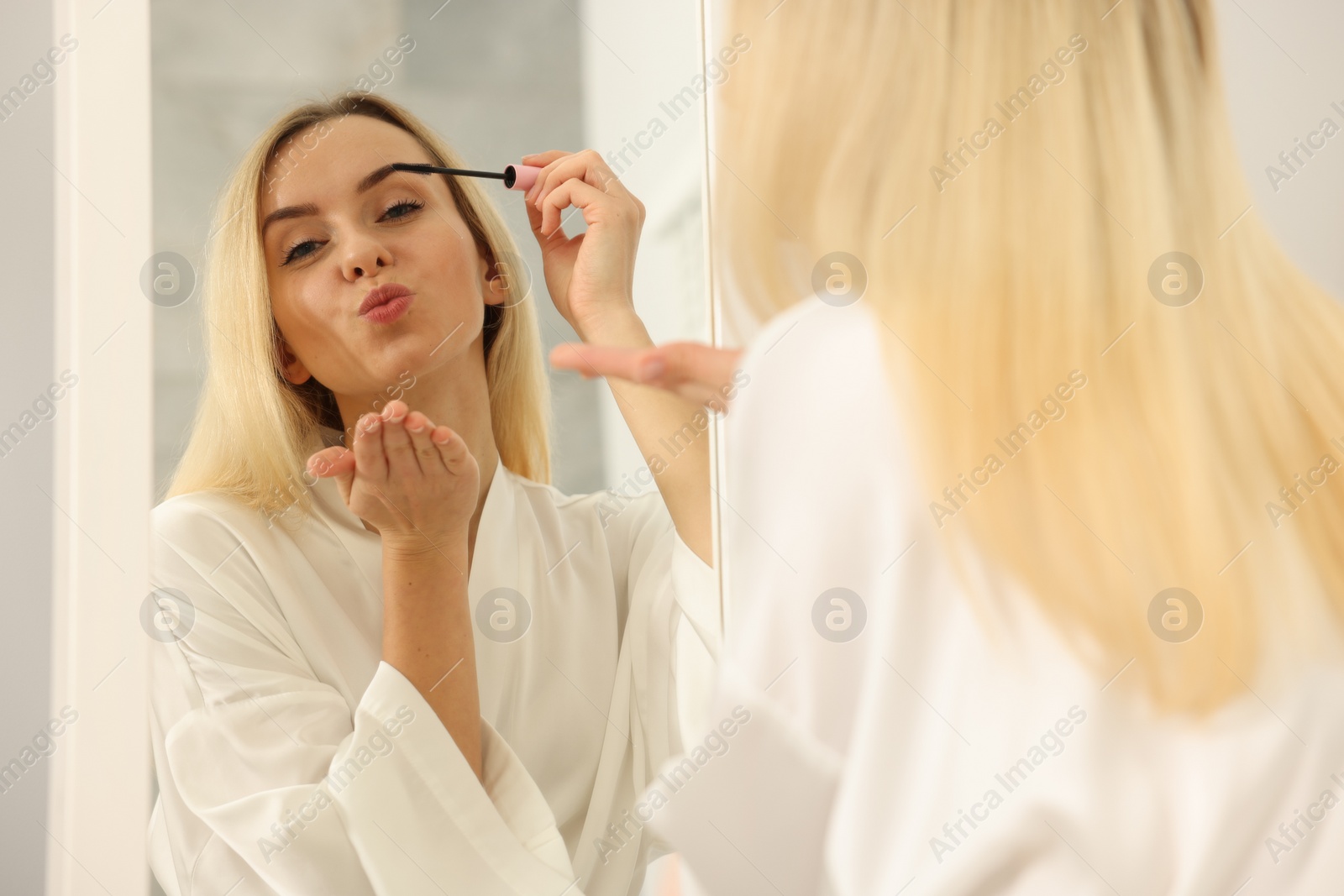 Photo of Beautiful woman blowing kiss while applying mascara near mirror in bathroom