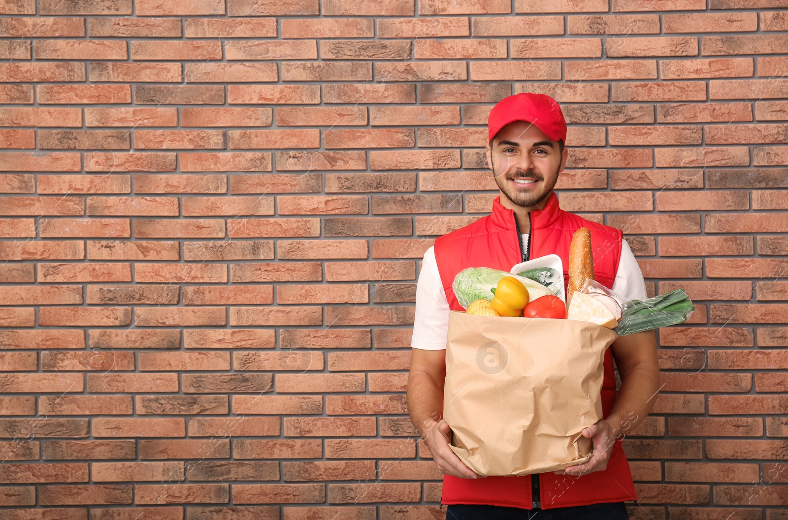 Photo of Food delivery courier holding paper bag with products near brick wall. Space for text