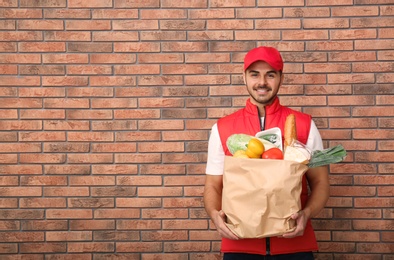 Photo of Food delivery courier holding paper bag with products near brick wall. Space for text