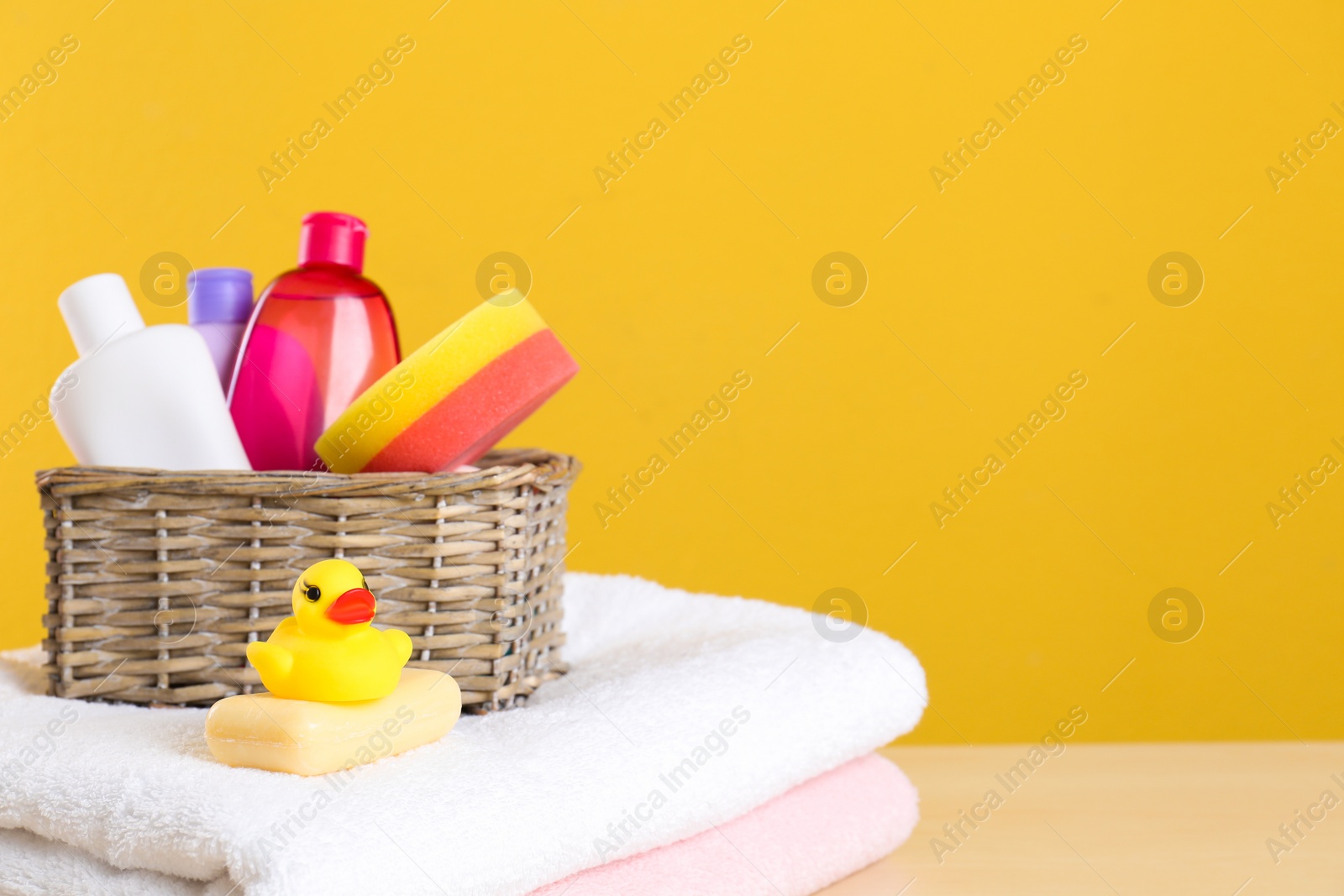 Photo of Wicker basket with baby cosmetic products, toy and towels on table against color background. Space for text