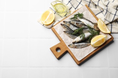 Photo of Fresh raw sprats, dill, oil and cut lemon on white tiled table, flat lay. Space for text