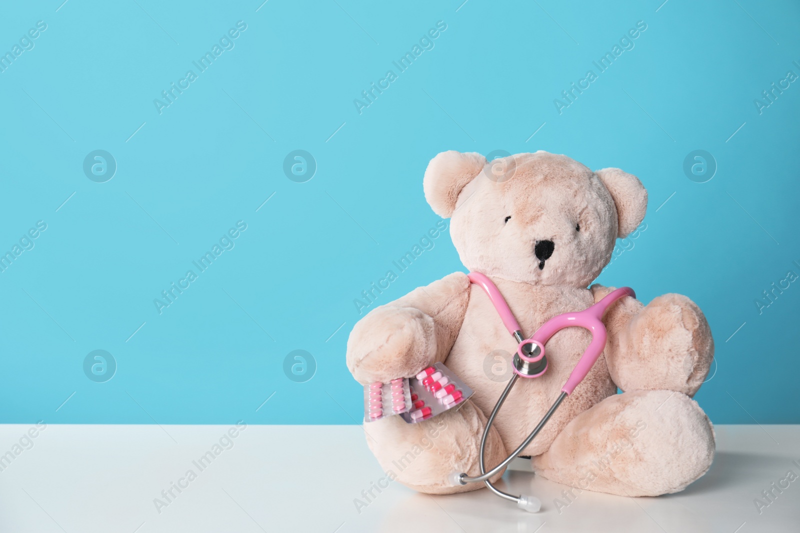 Photo of Toy bear with stethoscope and pills on table against color background, space for text. Children's hospital