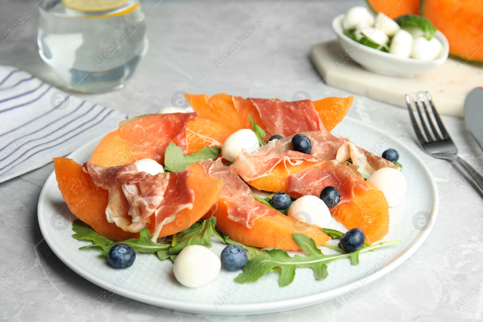 Photo of Fresh melon with prosciutto, mozzarella and arugula on light grey table