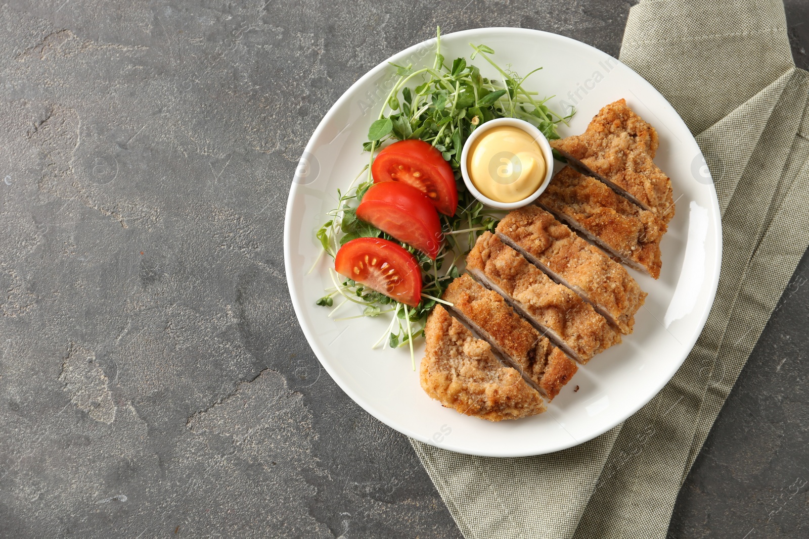 Photo of Tasty cut schnitzel served with sauce, microgreens and tomato on grey textured table, top view. Space for text
