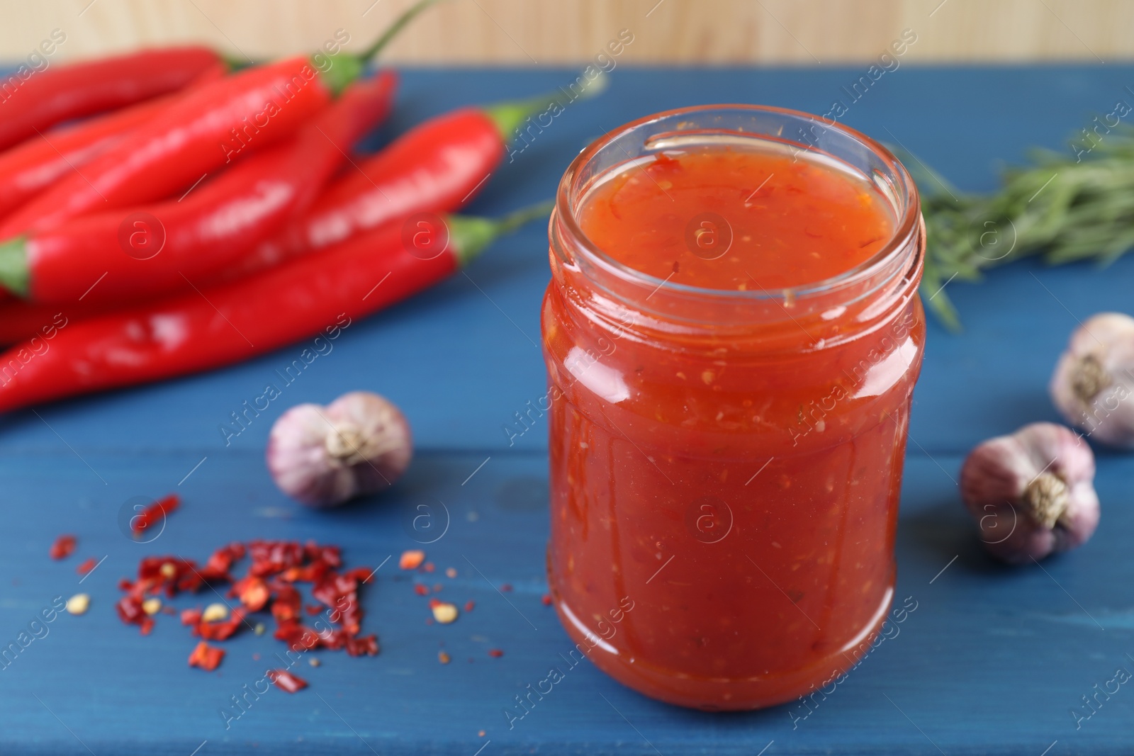 Photo of Spicy chili sauce in jar, garlic, peppers and rosemary on blue wooden table, closeup