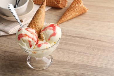 Photo of Delicious scoops of vanilla ice cream with wafer cone and strawberry topping in glass dessert bowl on wooden table, closeup. Space for text