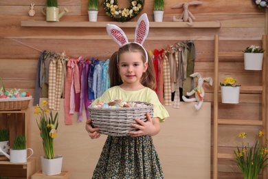 Photo of Adorable little girl with bunny ears and basket full of dyed eggs in Easter photo zone