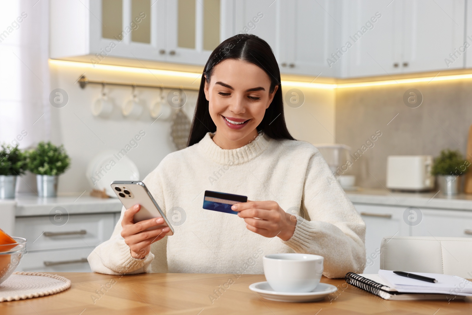 Photo of Happy young woman with smartphone and credit card shopping online at wooden table in kitchen