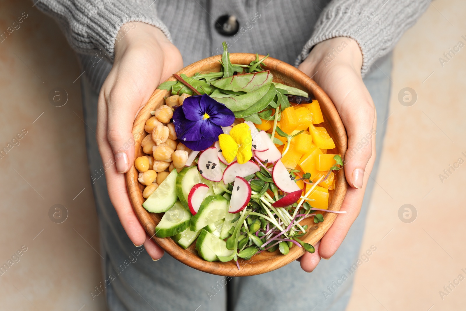 Photo of Woman holding delicious vegan bowl with cucumbers, chickpeas and violet flowers on beige background, closeup