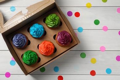 Box with different cupcakes and confetti on white wooden table, flat lay. Space for text