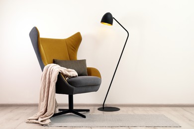 Photo of Comfortable armchair with blanket and floor lamp near white wall indoors