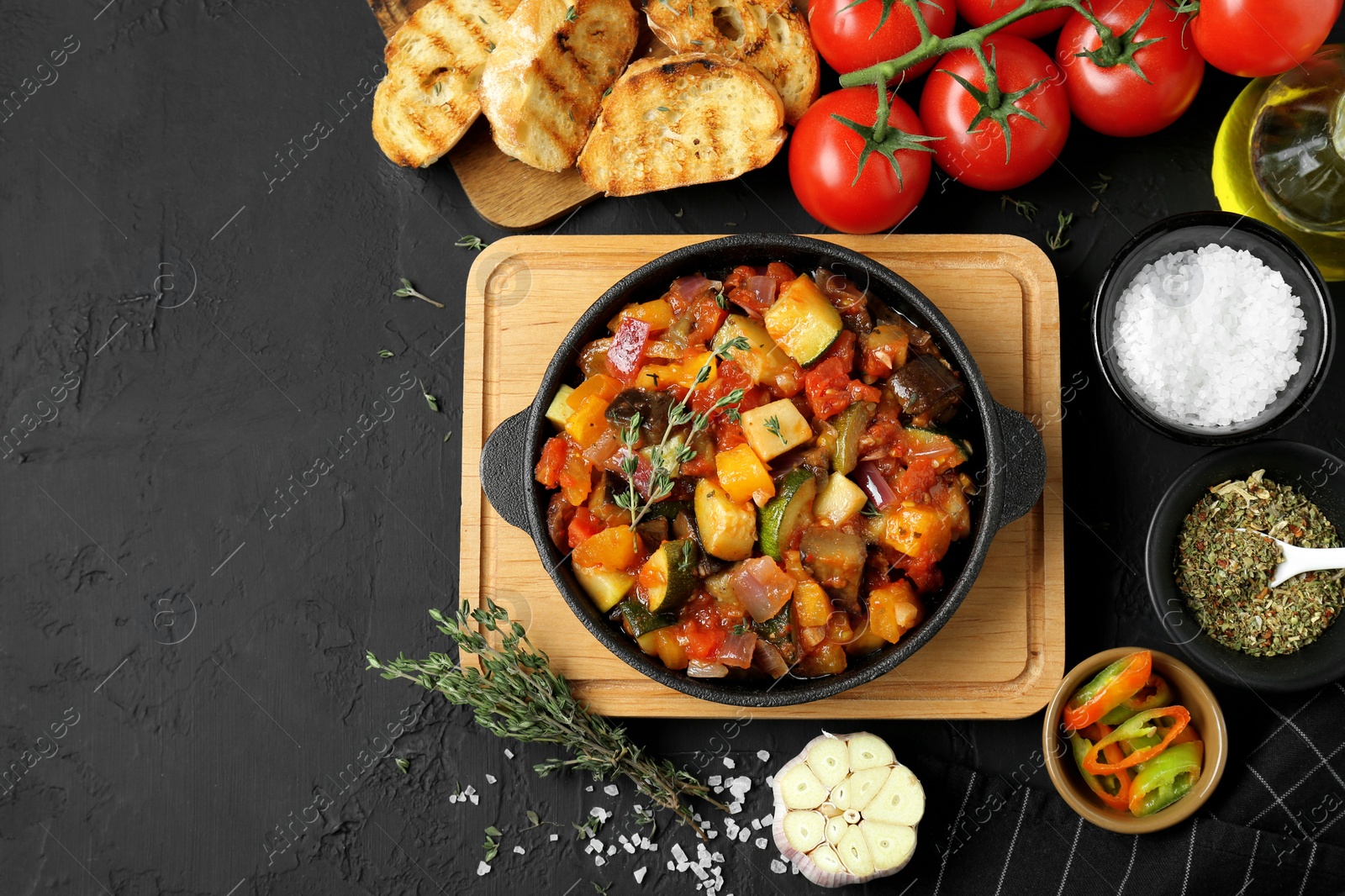 Photo of Dish with tasty ratatouille, ingredients and bread on black table, flat lay. Space for text