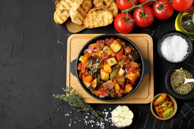 Photo of Dish with tasty ratatouille, ingredients and bread on black table, flat lay. Space for text