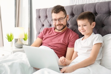 Photo of Little boy and his dad using laptop in bedroom