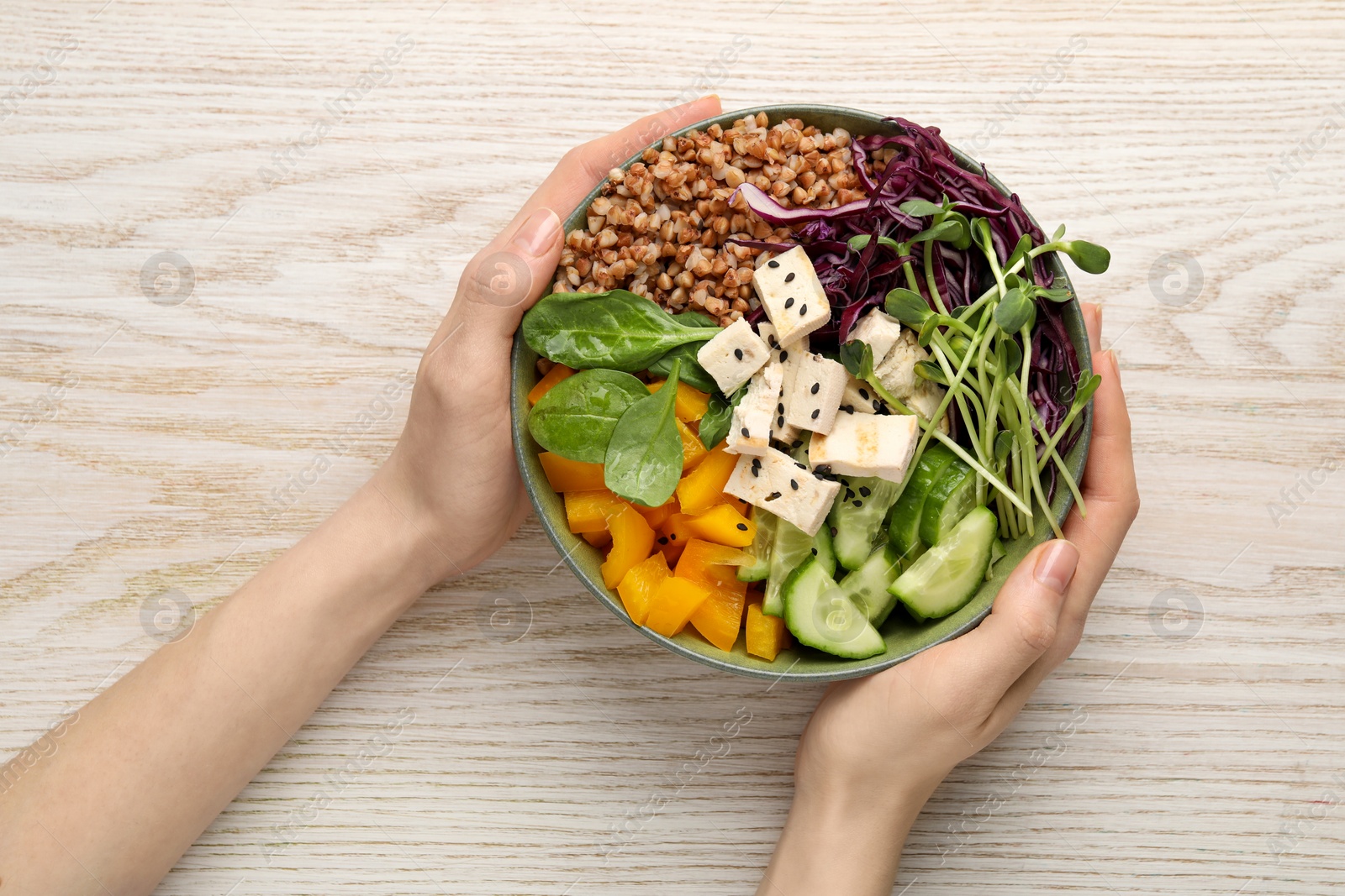 Photo of Woman holding delicious vegan bowl with tomatoes, microgreens and buckwheat at white wooden table, top view