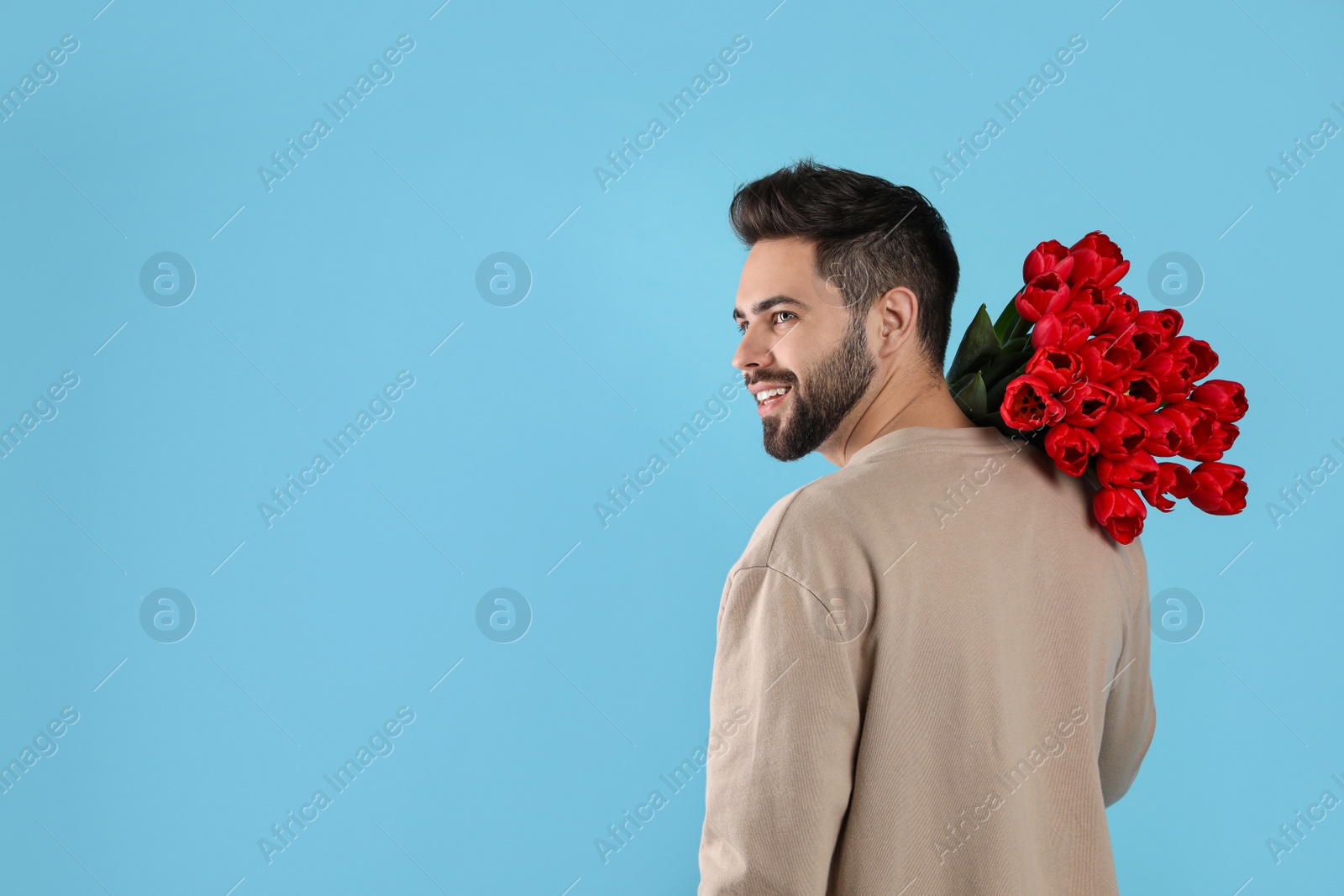 Photo of Happy man with red tulip bouquet on light blue background, space for text. 8th of March celebration