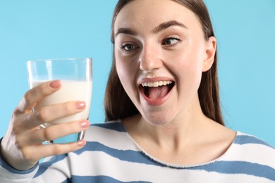 Emotional woman with milk mustache holding glass of tasty dairy drink on light blue background