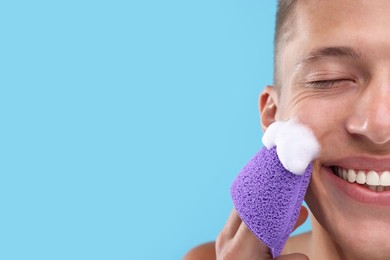 Photo of Happy young man washing his face with sponge on light blue background, closeup. Space for text