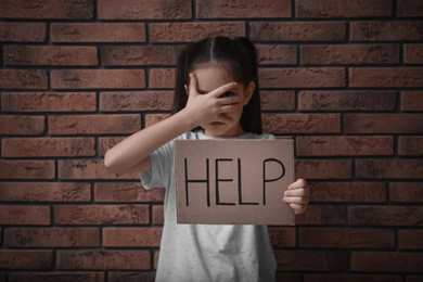 Photo of Sad little girl with sign HELP near brick wall. Child in danger